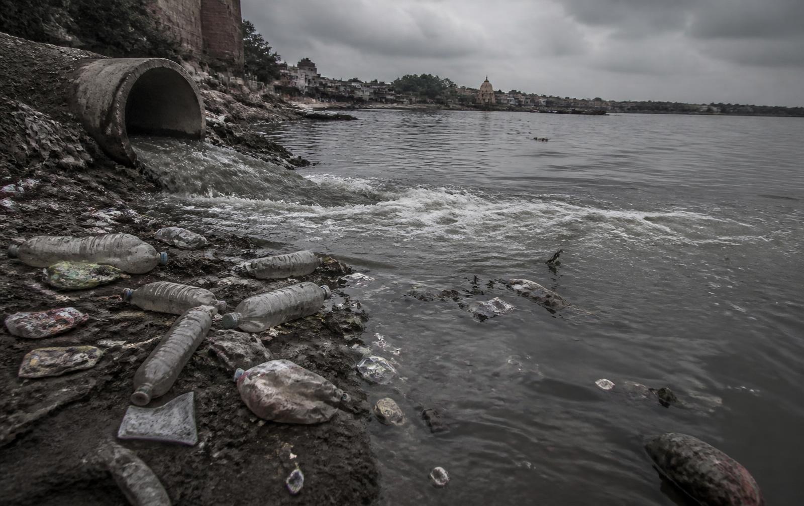 A drain outflow meeting an Indian river, with plastic bottles and packaging along the bank